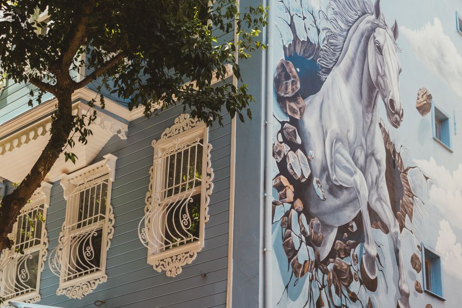 Artistic mural of a horse breaking through a wall on a blue house facade with ornate windows.