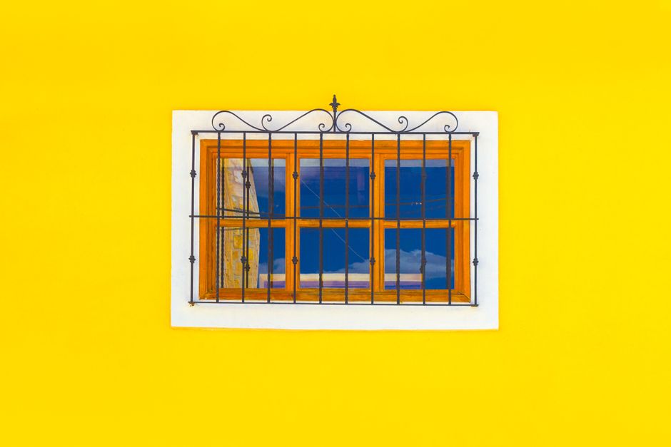Small horizontal window with wooden frame and metal bar placed on yellow wall of building in daytime in city outside